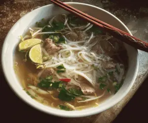 Steaming bowl of traditional Vietnamese pho with rice noodles, sliced beef, fresh herbs, lime, and chili peppers